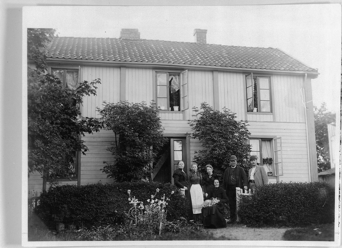 Johs. (Johannes) Johannessen, EKSTERIØR, STRANDSTUEN, KIRKEGATA. 11, FAMILIEN FLAGSTAD FORAN HUSET. NR. 2 FRA HØYRE KORPSBØRSEMAKER O. M. FLAGSTAD, MED SØNNEN KAPELLMESTER M. FLAGSTAD OG HUSTRU PIANISTINNEN M. FLAGSTAD NIELSEN PÅ HVER SIDE. YTTERST TIL VENSTRE GAMLE BESTEMOR OLIA FØDT PÅ VALSIG I FURNES SITTER I STOLEN MED KATT…