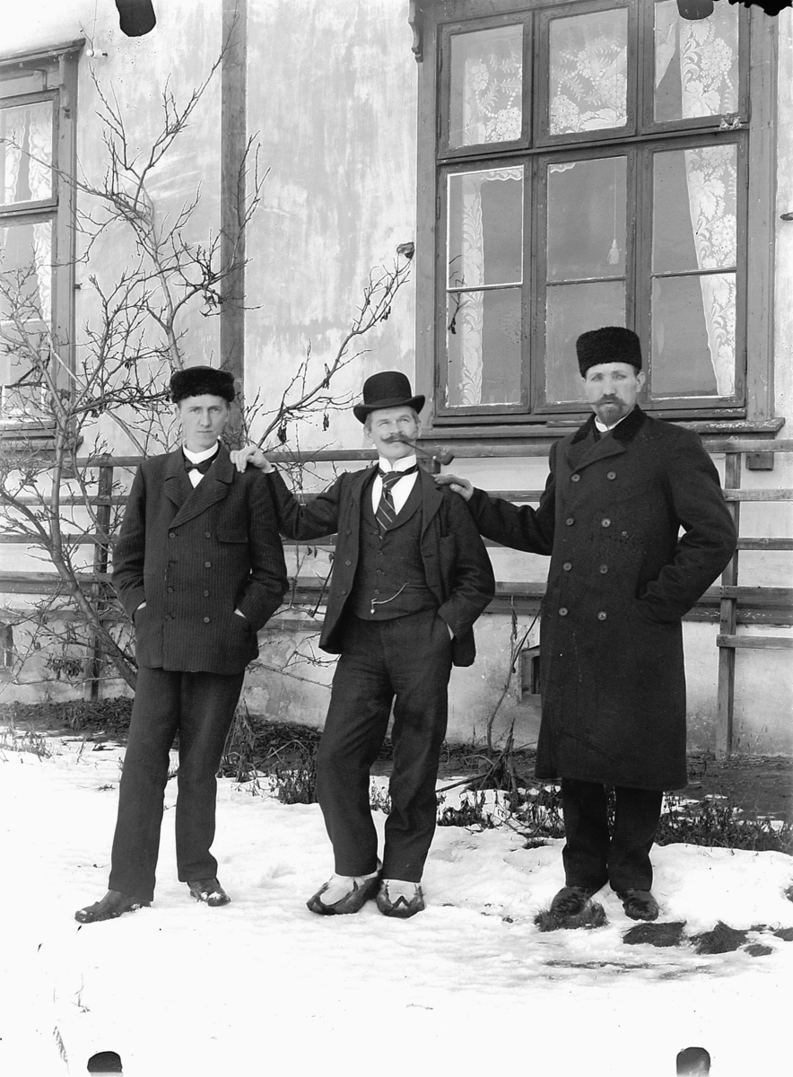 Ragnvald Andersen, Vinterbilde, tre menn i dress foran hus som er rappet. Mannen i midten røyker pipe. Ukjente.