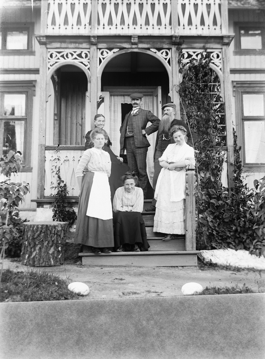 Ragnvald Andersen, Gr. 6. Familien Danielsen. To menn og fire kvinner foran veranda. Sveitserhus med hage. Kjøpmann Danielsen, Lundberg, Haraldstad, Furnesvegen. Fotografens svigerfamilie, han sjøl i midten? Fru Pernille Danielsen, F. S. Fokstuen, kjøpmann Danielsen, Olga Andersen og Dagmar Danielsen, døtre av kjøpma…