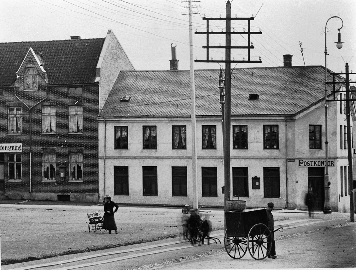 Hans Emil Erichsen, Østre Torg i Hamar med bebyggelsen langs Vangsvegen. Hamar Postkontor. Mann med håndkjerre. Lysstolper og gatebelysning. Utsnitt.