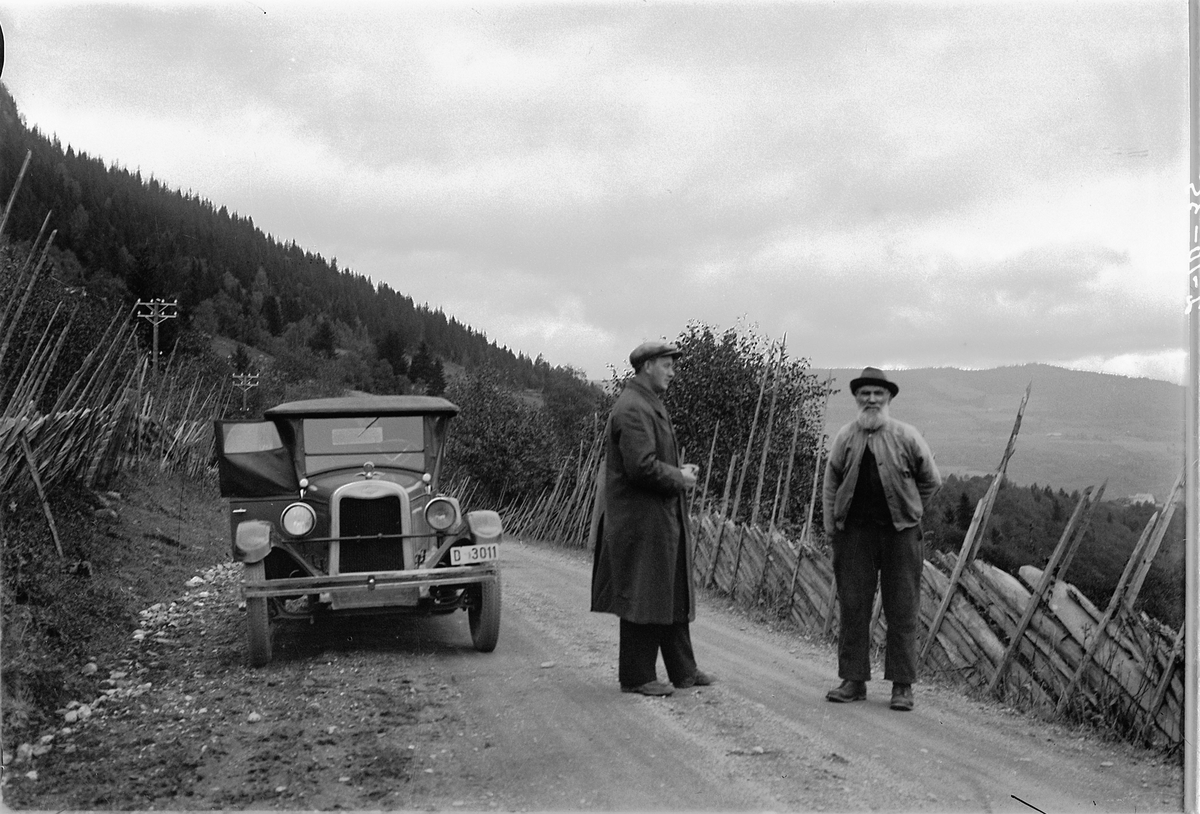 Simen Baardseth, Brøttumsbakkene i Ringsaker. Lærer Karl Tallhang t. v. den andre er ukjent, foran en bil med nummer D-3011, en Chevrolet 1925-26. Grusveg, skigard, telefonstolper I Bilboken for Norge 1930 står registreringsnummeret på en Chevrolet personbil tilh. L. O. Lund, Vang. I Norges Bilbok 1935 på ditto 192…