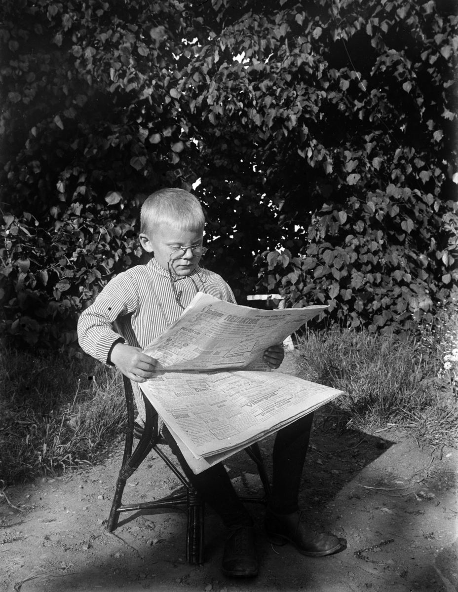 Kristian Heramb, Eksteriør, hage. En gutt sitter i en stol og leser avisen.