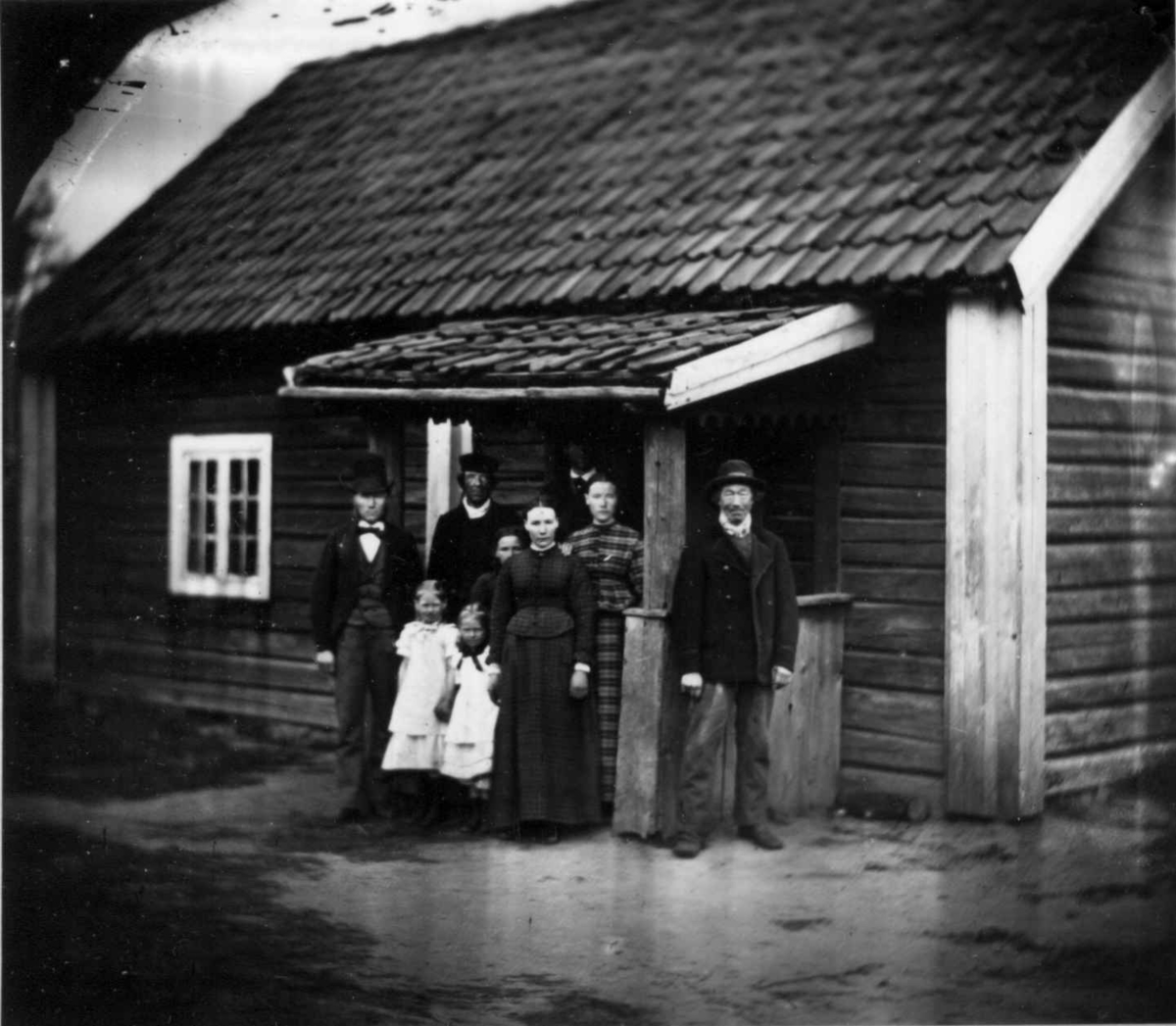 Fredrik Emil Faye, Husmannsfamilien Baskop, Dal gård, Ullensaker. Gruppe poserer foran inngangsparti til bolighus, ca. 1885. Fra portrettserie av personer som bodde på eller besøkte Dal gård, Ullensaker, fotografert av gårdens eier, kammerherre Fredrik Emil Faye (1844-1903) i årene 1875-1900.