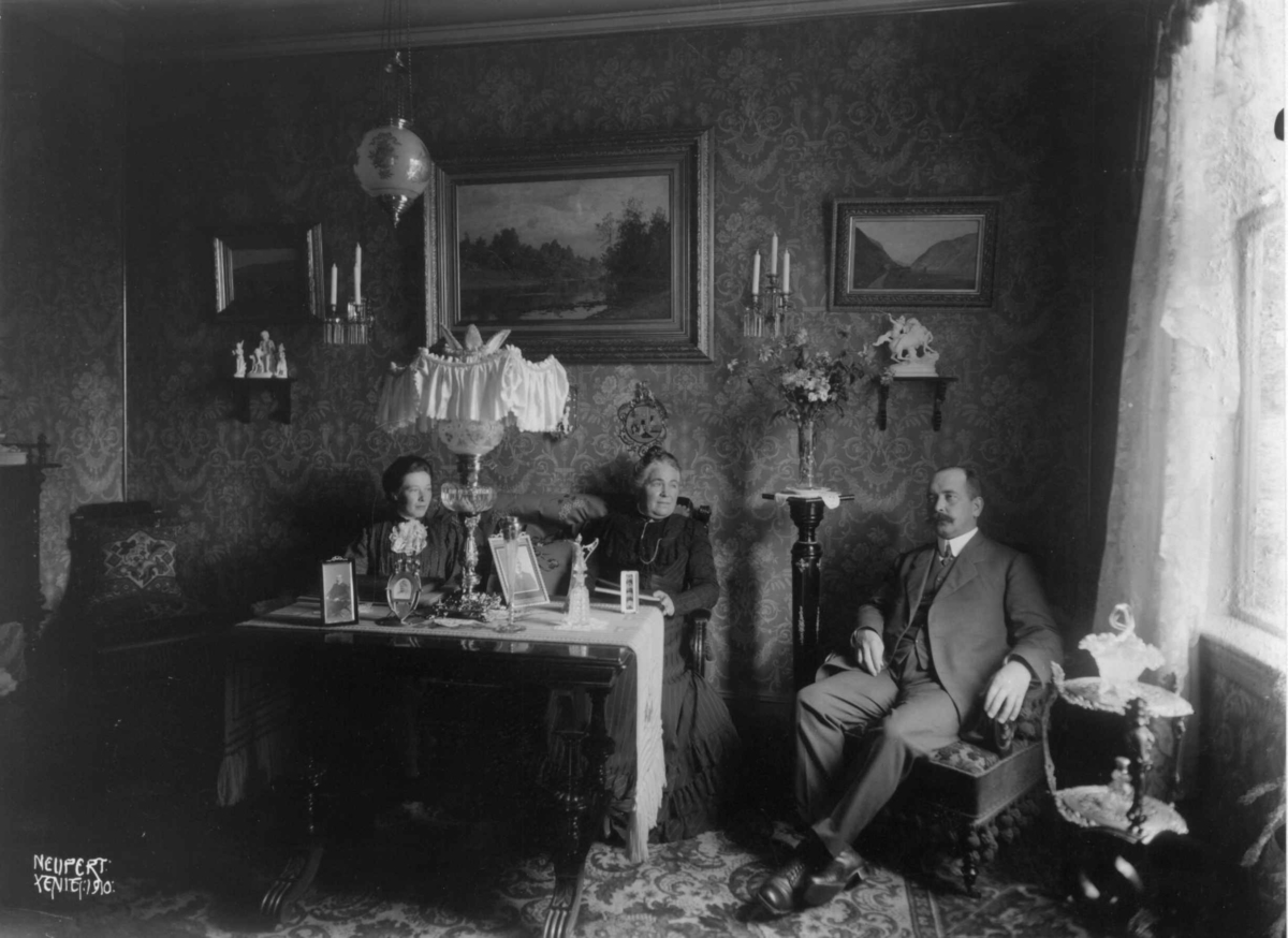 Hermann Christian Neupert, Stueinteriør. Holm. 1908-1910. To kvinner og en mann sitter rundt et bord. Malerier på veggene..