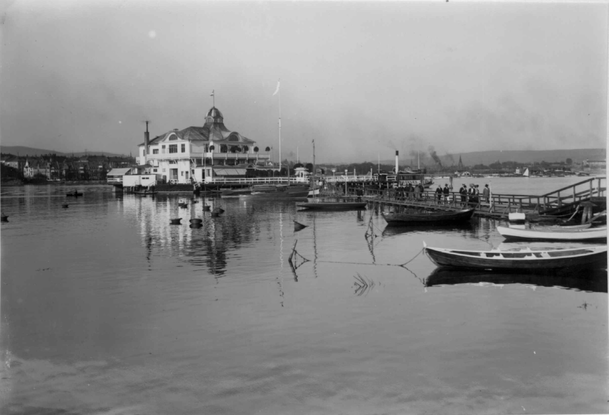 Hermann Christian Neupert, Oslo, Dronningen 1909. Bygningen, båter, ferge og mennesker på brua. Sett fra vannet.