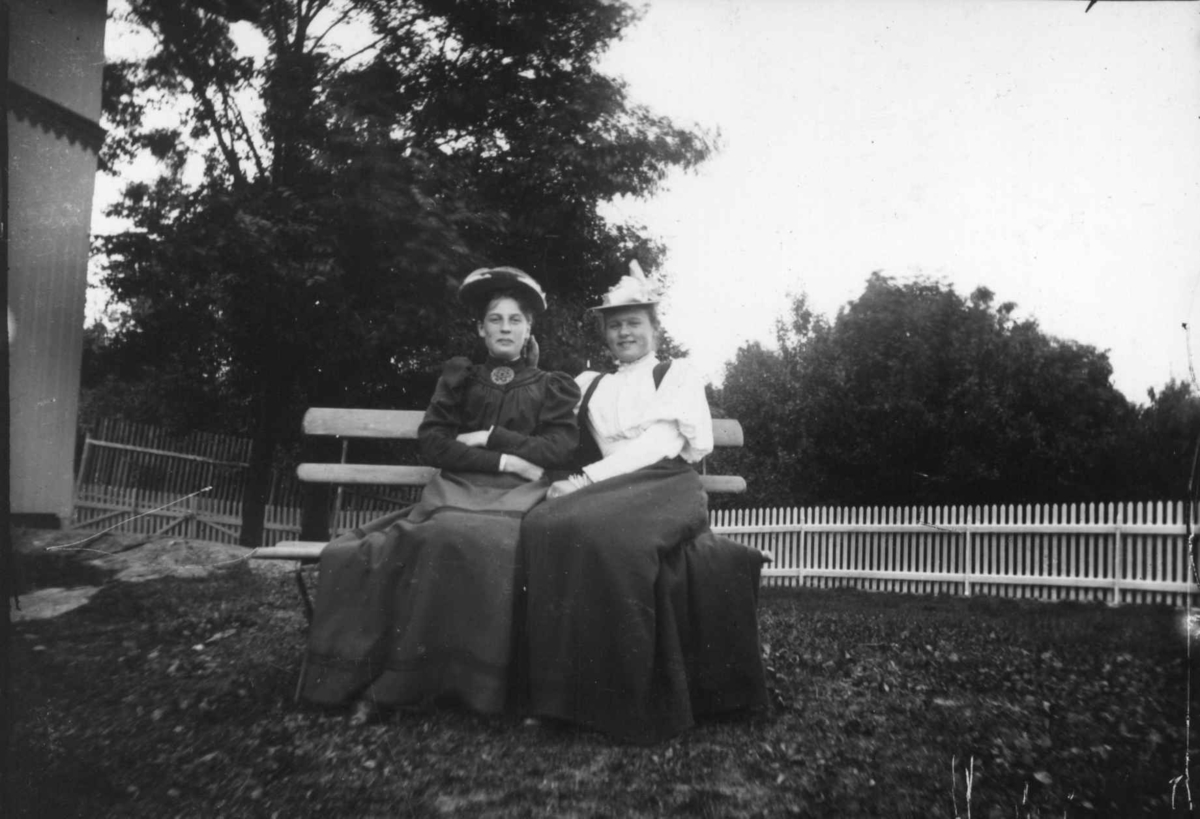 Hermann Christian Neupert, Drakt 1908-1910. To kvinner sittende på en benk i en hage. Bærer hatter.