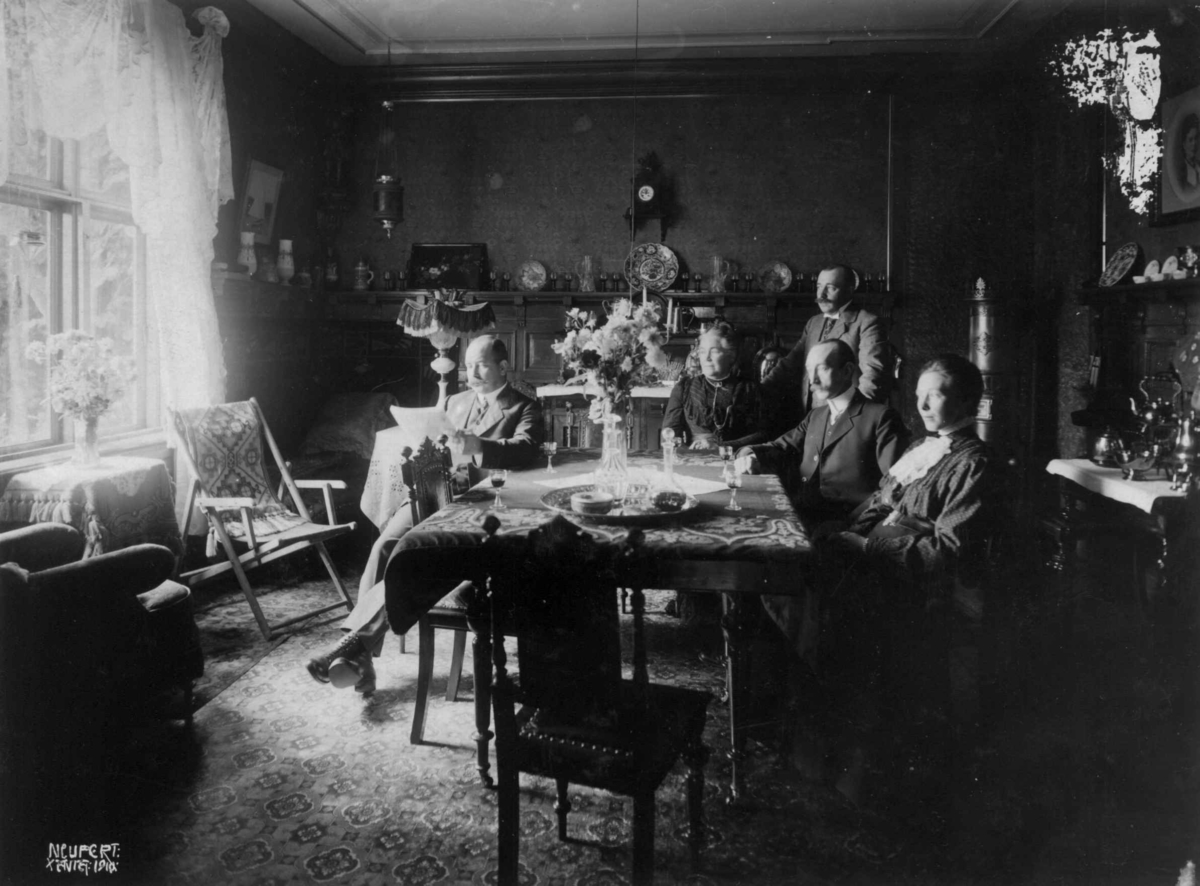Hermann Christian Neupert, Holm. 1910. Stueinteriør. Menn og kvinner rundt et bord. På bordet står det glass og karafler.