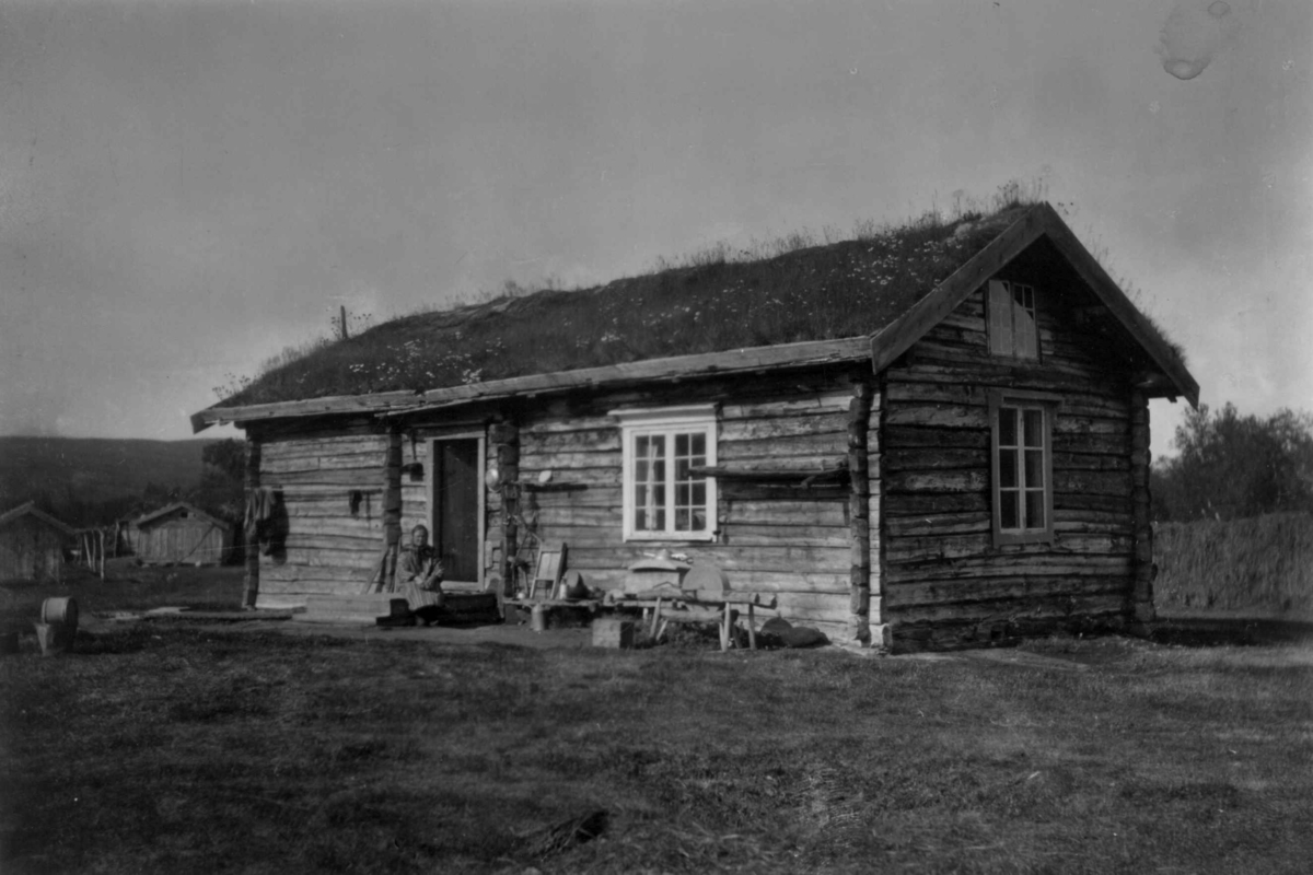 Konrad Nielsen, Stue eller bolig i laftet tømmer og med torvtak, uthusbygninger i bakgrunnen. En kvinne sitter på trappen til inngangsøren. Alleknjarg 1899.