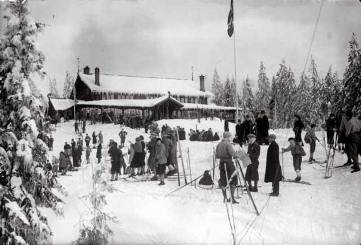 Hermann Christian Neupert, Frognerseteren Restaurant, Nordmarka, Oslo. Vintermotiv. Skiløpere foran restauranten. Flaggstand med flagg.