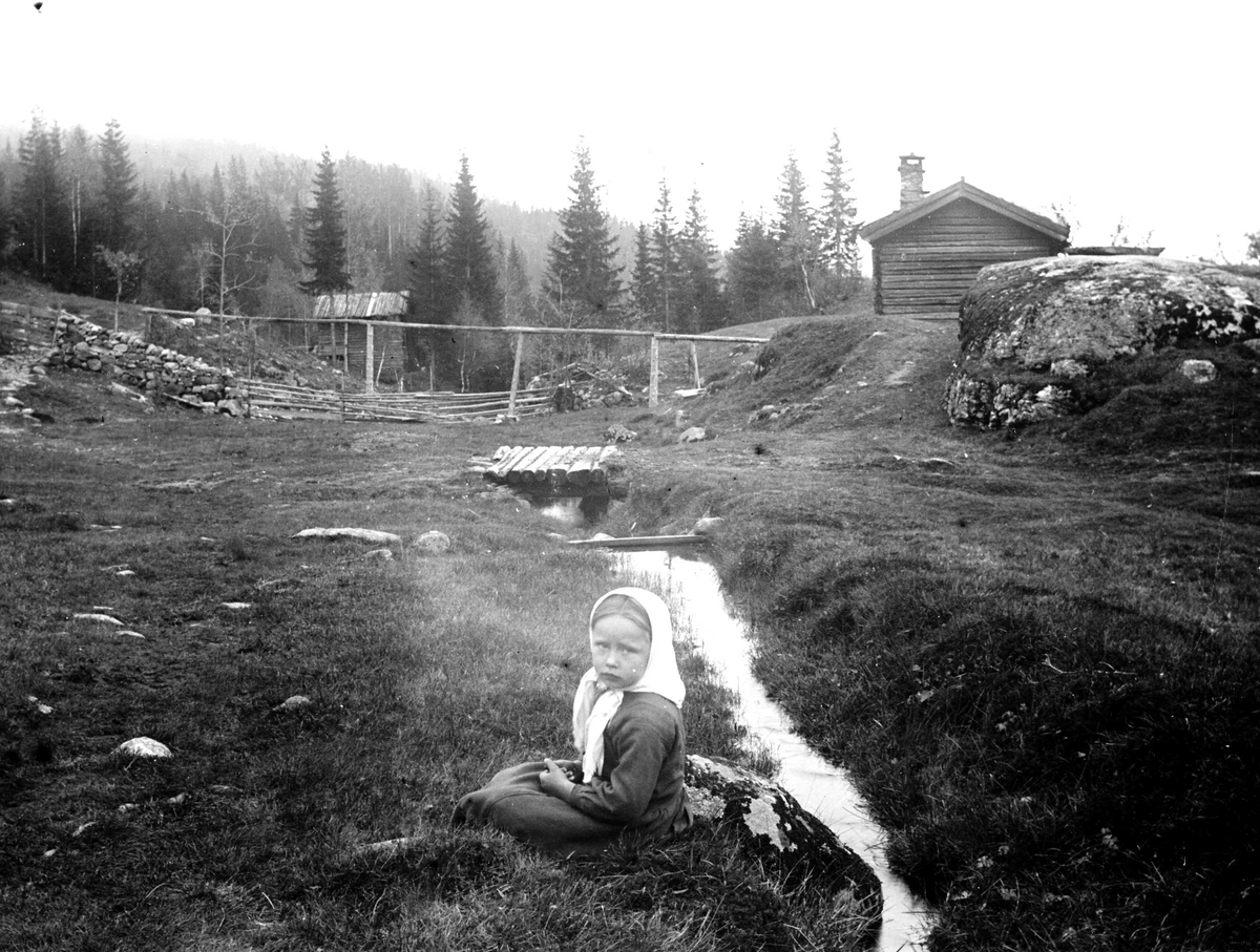 Juliane Fredrikke Langberg, Fra Eggedal, Sigdal, på besøk hos Th. Kittelsen. Fra beite(?)område ved gårdsbebyggelse med div. gjerder. Lite elveløp med bro av tømmerstokker. Jente med hvitt skaut sitter i forgrunnen. Fra malerinne Juliane Fredrikke Langbergs samling, tatt 1895-1910 med motivkrets slekt og venner samt steder ti…