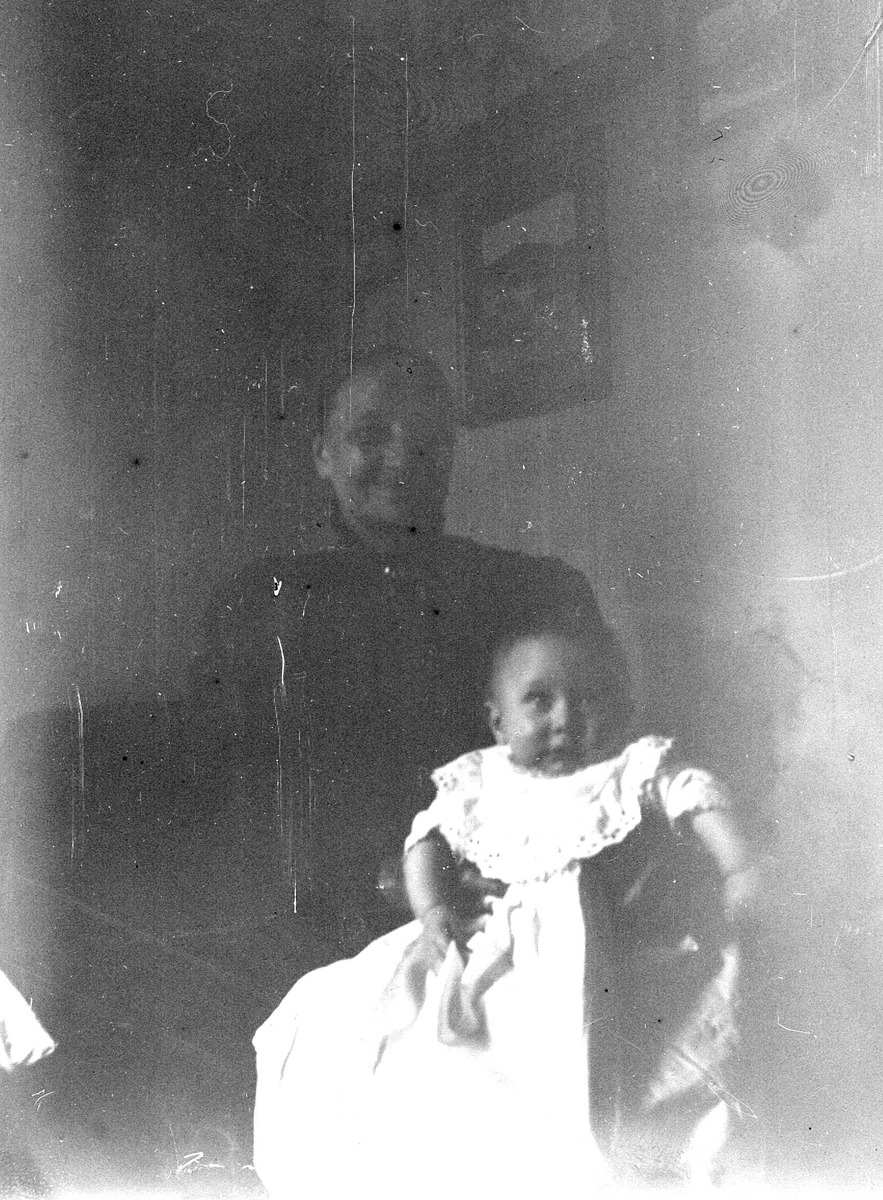 Juliane Fredrikke Langberg, Dåpsbarn i dåpskjole på fanget til en eldre kvinne i interiør (mørk bakgrunn).Ukjente.Noe uskarpt bilde. Fra malerinne Juliane Fredrikke Langbergs (1856-1930) samling, tatt ca.1895-1910 med motivkrets slekt og venner samt steder tilknyttet samvær med disse.