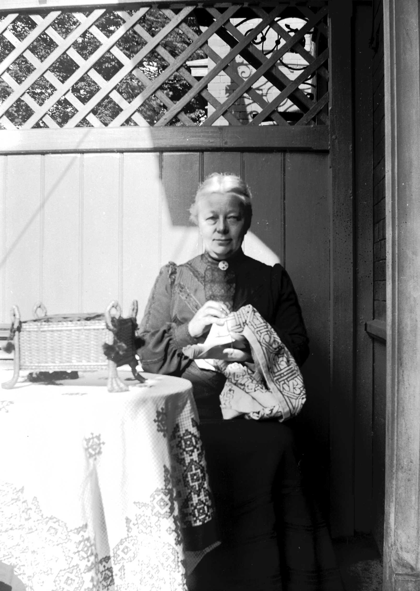 Juliane Fredrikke Langberg, Portrett av eldre kvinne som broderer utendørs, i hagestue(?). På bordet med brodert duk står en sykurv(?) på bein. Muligens på kappelangården Bergenhus i Rakkestad eller prestegården i Hurum, hvor fotografens søster Louise var gift med presten Olaf A.Holm. Fra malerinne Juliane Fredrikke Langbergs…