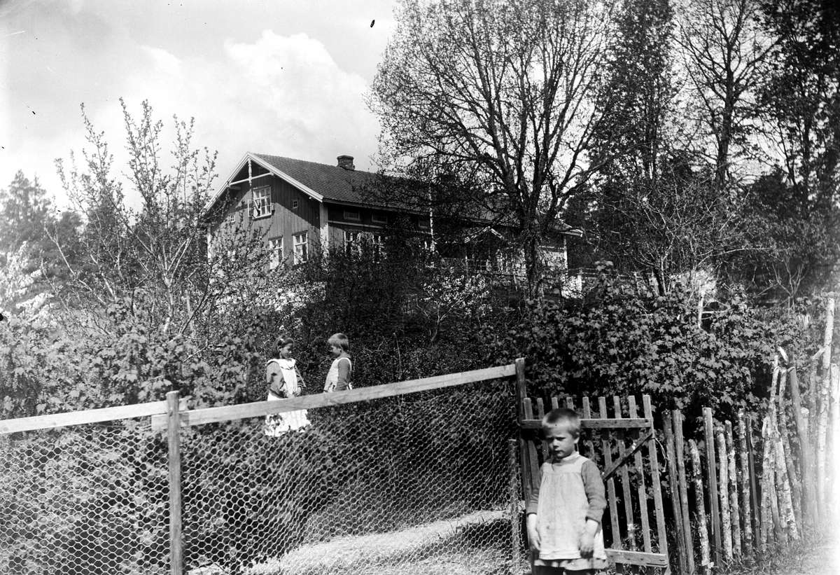 Juliane Fredrikke Langberg, Kapellangården Bergenhus i Rakkestad med 3 av prestebarna i hagen foran. I forgrunnen utenfor gjerde med port, Christian L. Holm, inne i hagen Emily og Olaf L. Holm. Holm-barna er barn av sokneprest i Rakkestad 1884-97, Olaf A. Holm, g. med fotografens søster Louise. Fra malerinne Juliane Fredrikke…
