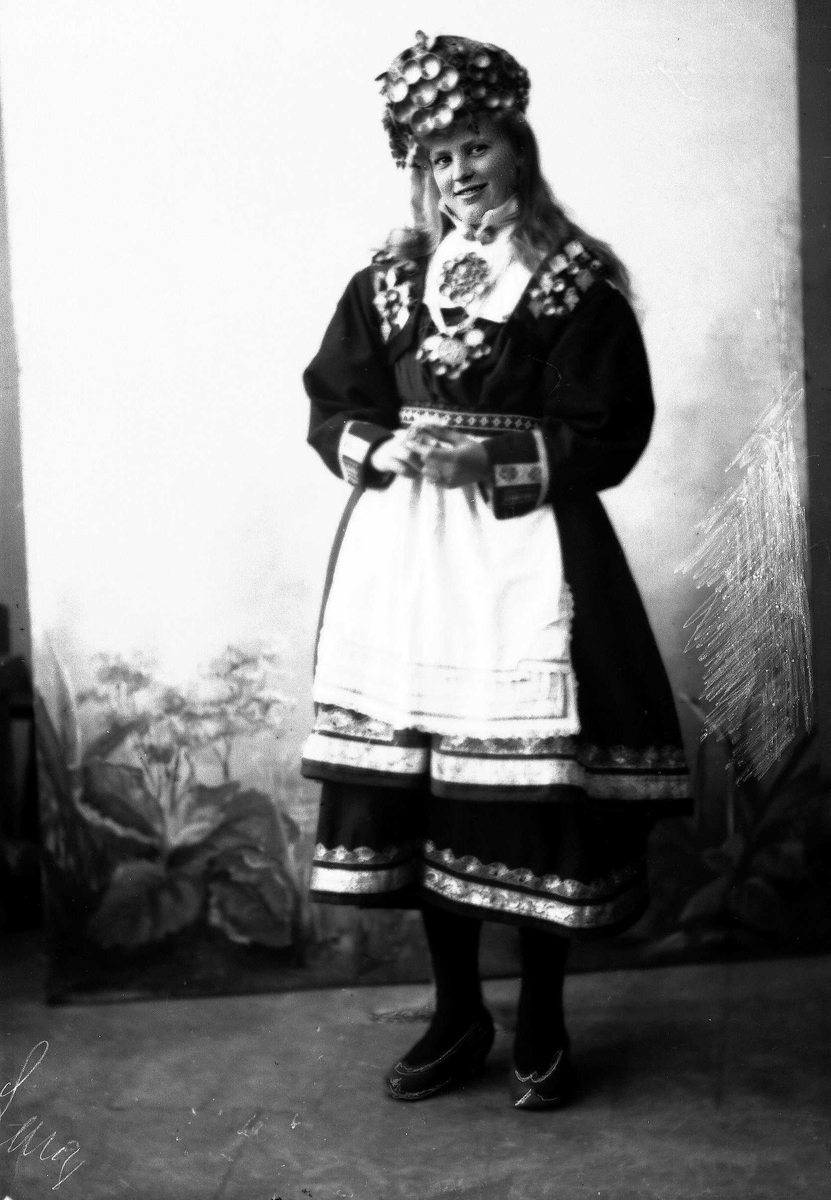 Solveig Lund, Studiofotografi av kvinne med brudedrakt og brudehatt/krone på hodet. Stående foran malt bakgrunn med blomster. Helfigur Frontalt vendt. Ser ut av bildet.