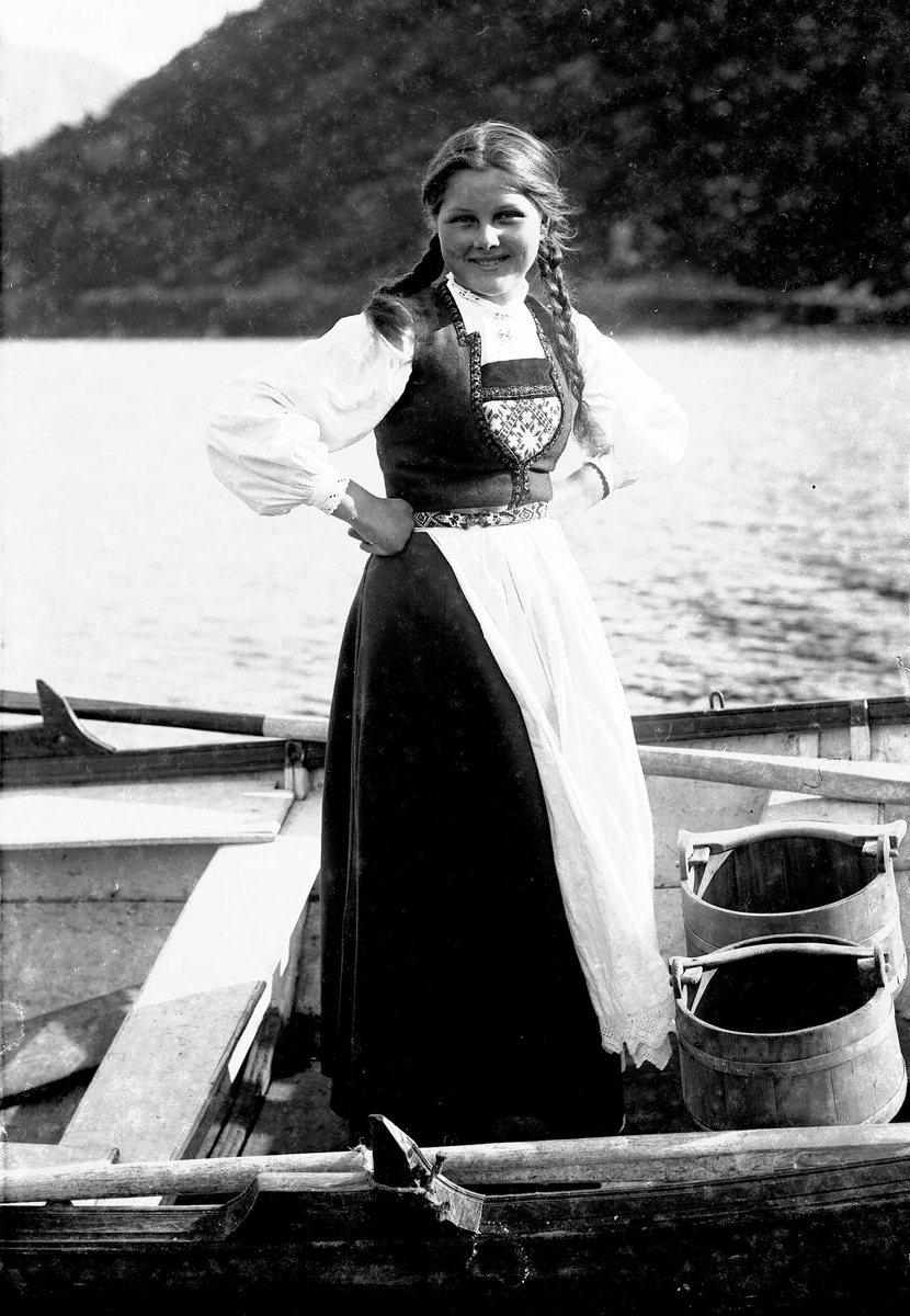 Solveig Lund, Jente i Hardangerdrakt står i robåt med vannbøtter.