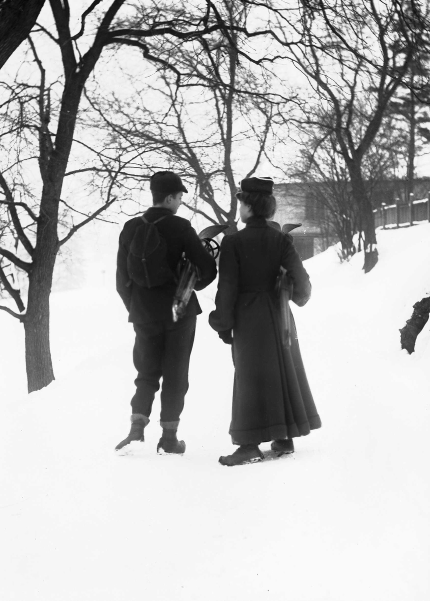 Solveig Lund, Mann og kvinne i snødekt landskap. Fotografert bakfra - begge med ski under armen og mannen med sekk på ryggen.