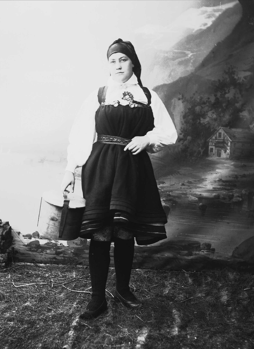 Solveig Lund, Studioportrett av kvinne i nasjonaldrakt som holder tine.Ser mot venstre i bildet. Bakgrunnen er malt med fjord og fjell landskap.