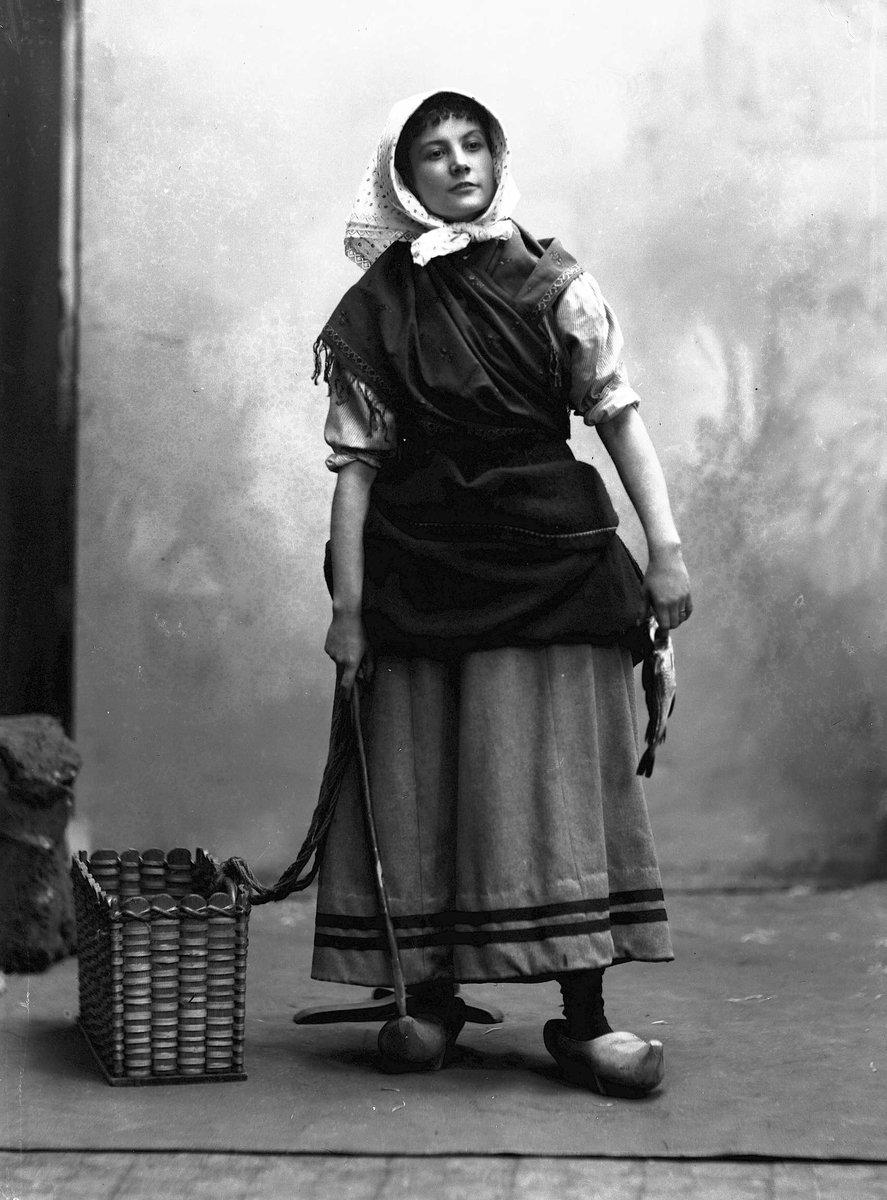 Solveig Lund, Studioportrett av kvinne i drakt med skaut på hodet. Hun med en fisk i den ene hånden og et redskap i den andre. På gulvet ved siden av henne står en trekutv. Bakgrunnen er malt. Gulvet er av tre med teppe på.