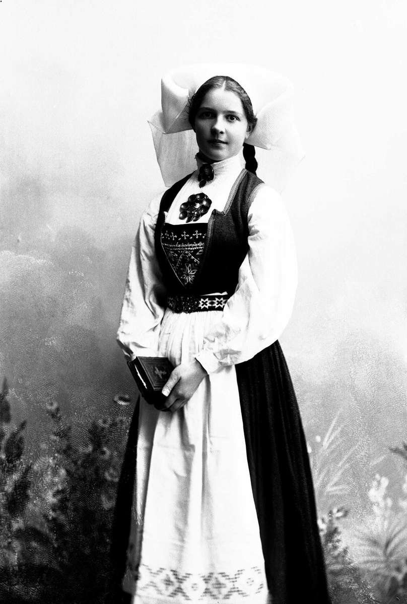 Solveig Lund, Studioportrett av kvinne i drakt med hodeplagg. Holder bibel/salmebok i hendene. Bakgrunnen er malt med blomster.