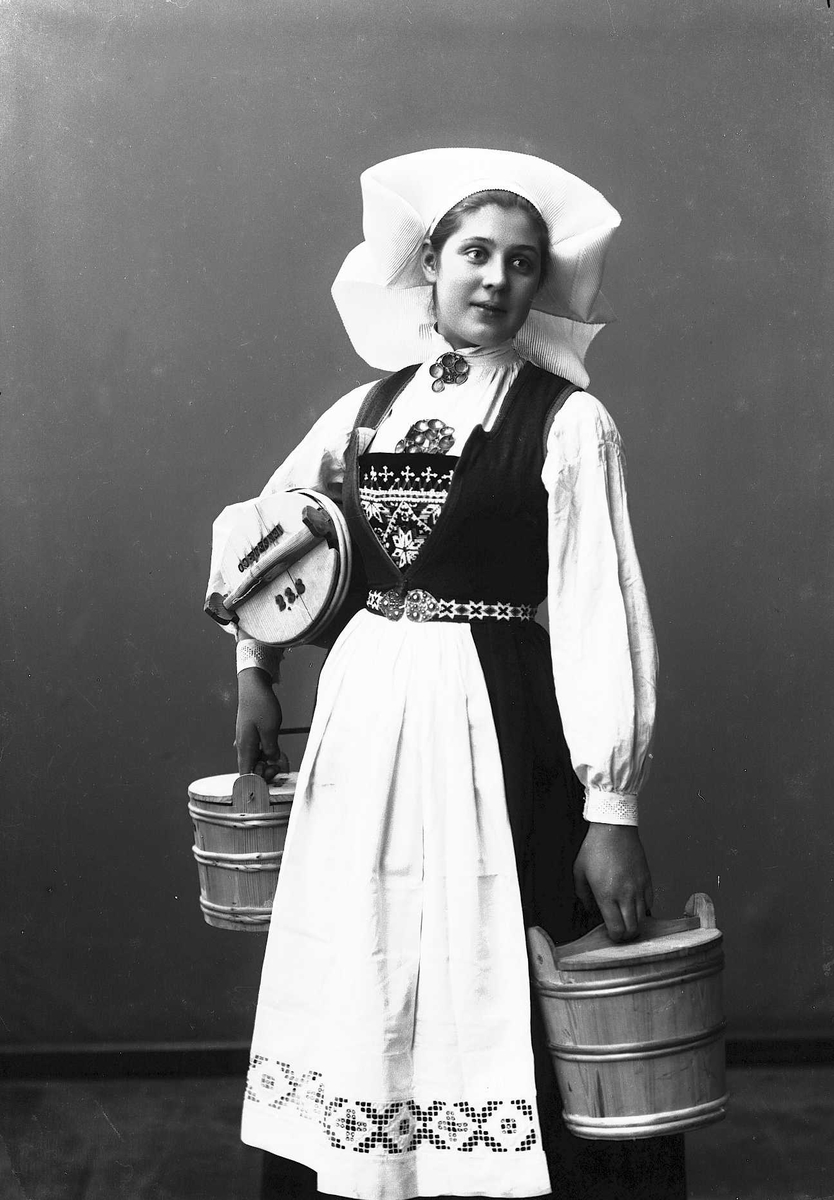 Solveig Lund, 3/4 portrett av kvinne i drakt og skaut, tatt i fotografens studio. Hun holder en tine i hver hånd og en under den høyre armen. Nøytral bakgrunn.