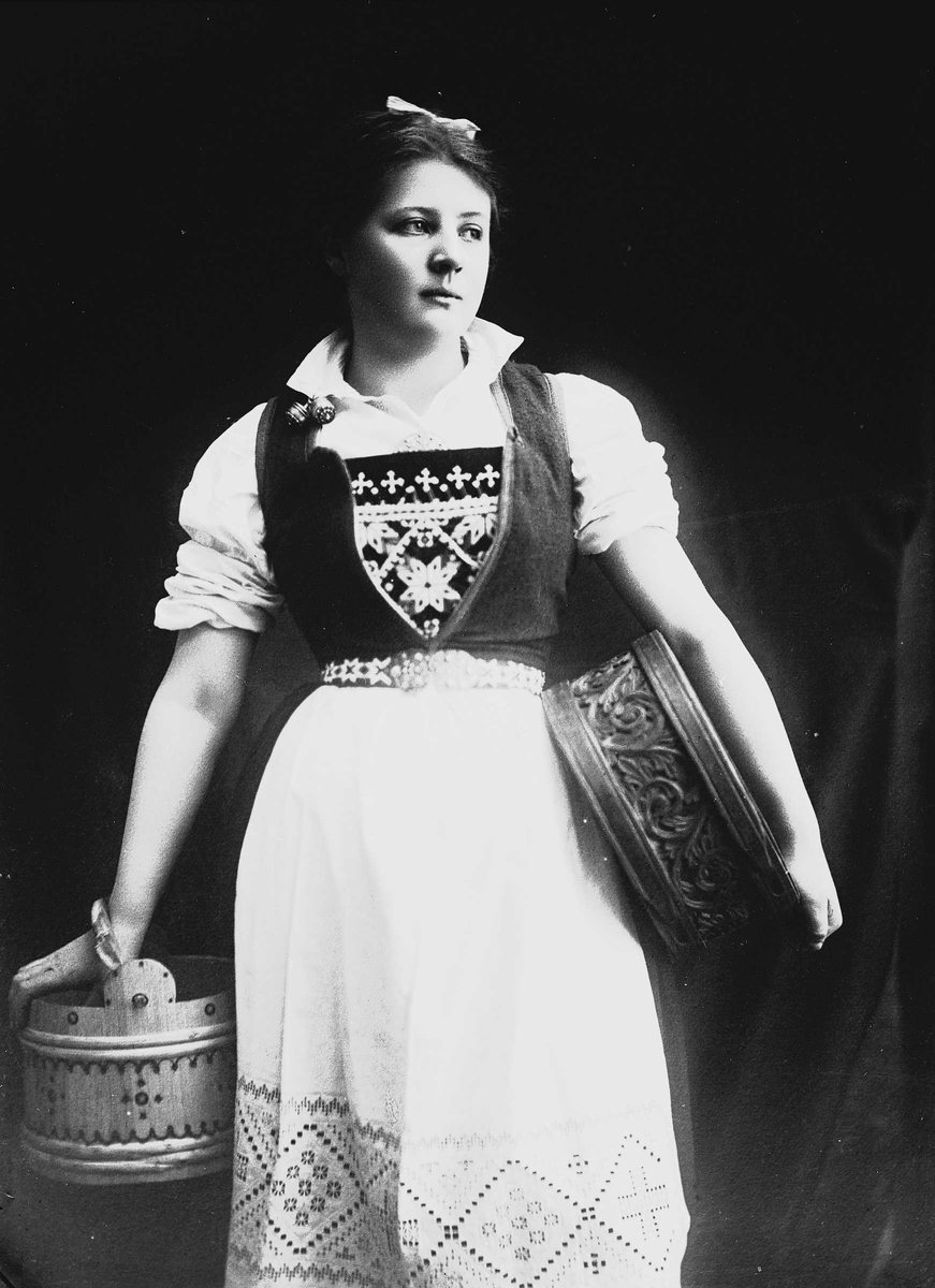 Solveig Lund, 3/4 portrett av kvinne i drakt med opprullede armer, tatt i fotografens studio. Hun holder en eske under den venstre armen og i den høyre en bøtte. Hun ser ut mot høyre i bildet. Nøytral bakgrunn.