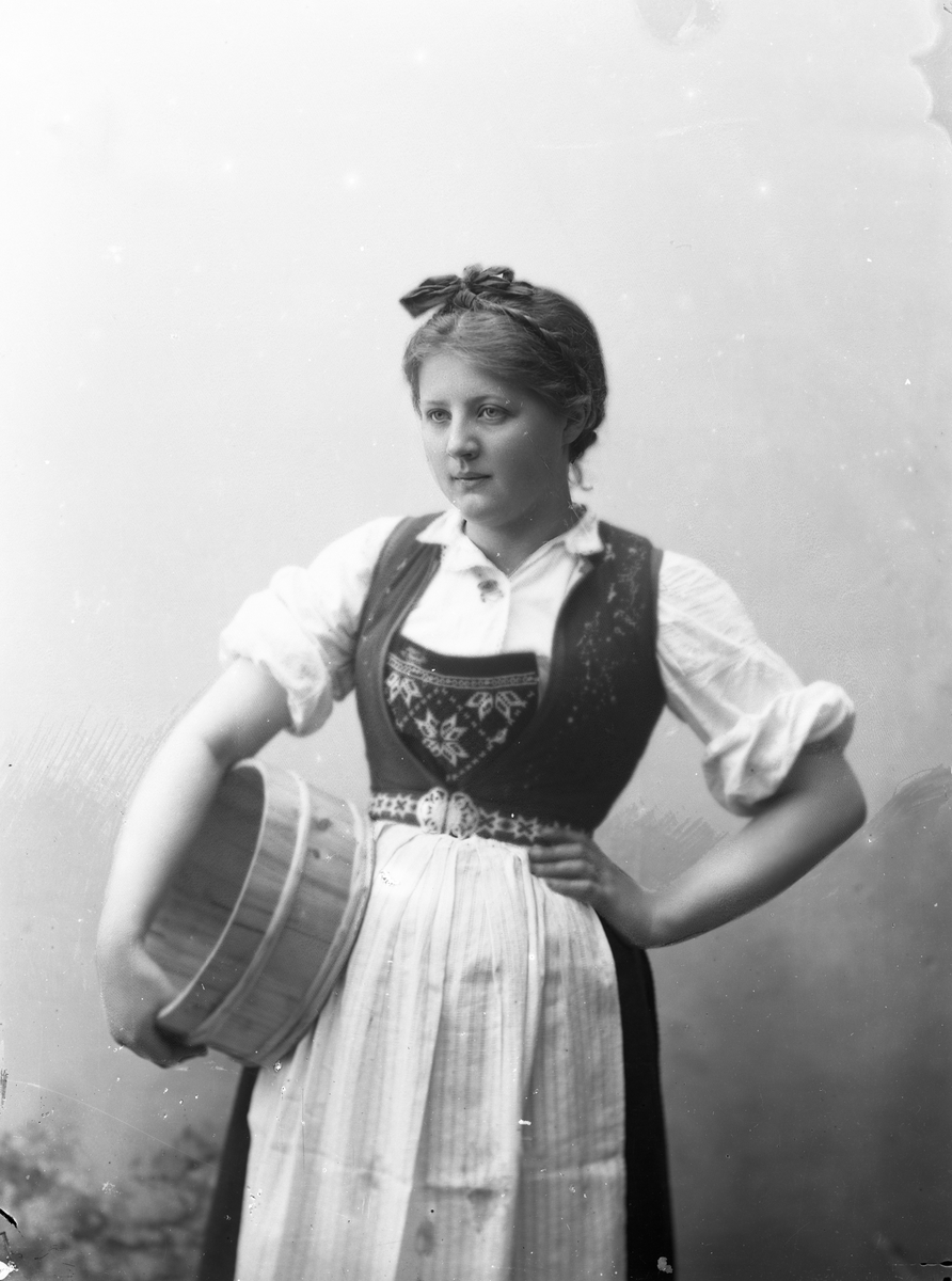 Solveig Lund, 3/4 portrett av kvinne i drakt med opprullede armer, tatt i fotografens studio. Hun holder en melkebunke under den høyre armen og den venstre på hoften. Hun ser ut mot venstre i bildet. Nøytral bakgrunn.