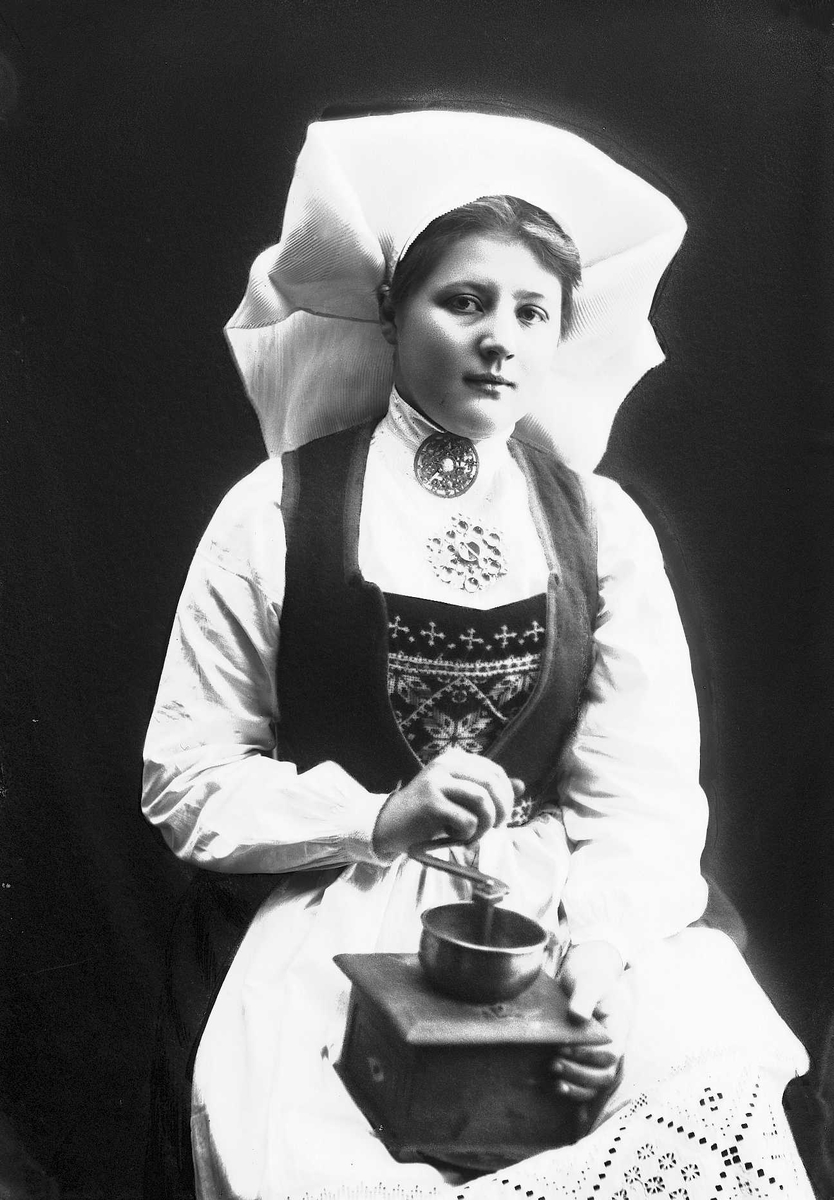 Solveig Lund, 1/2 portrett av kvinne i drakt og skaut, tatt i fotografens studio. Hun sitter og holder en kaffekvern på fanget. Nøytral bakgrunn.