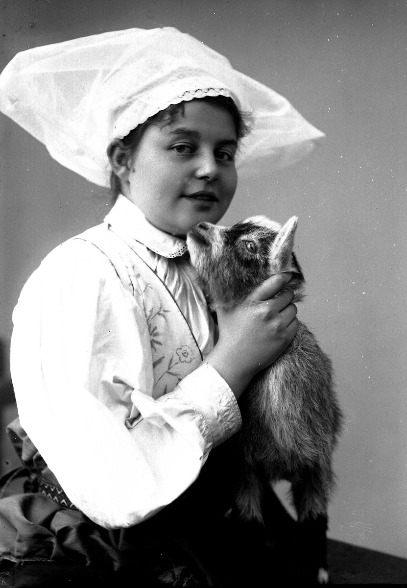 Solveig Lund, 1/2 portrett av kvinne i drakt og skaut, tatt i fotografens studio. Hun sitter med høyre side vendt mot fotografen og kikker ut på betrakteren. Hun holder en geit på fanget. Nøytral bakgrunn.