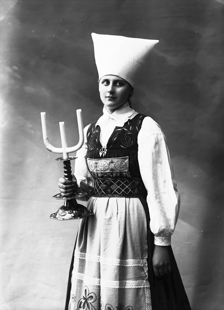 Solveig Lund, 3/4 dels portrett av kvinne i drakt med skaut/hatt på hodet, fotografert i studio. Hun holder i en trearmet lysestake i høyre hånd. Kvinnen ser ut mot høyre i fotografiet. Nøytral bakgrunn.