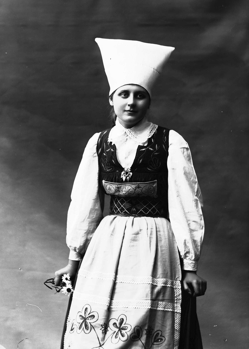 Solveig Lund, 3/4 del studioportrett av kvinne i drakt og skaut/hatt. Hun støtter seg mot noe vi ikke ser og i høyre hånd holder hun en prestekrage. Hun ser ut av fotografiet. Nøytral bakgrunn.