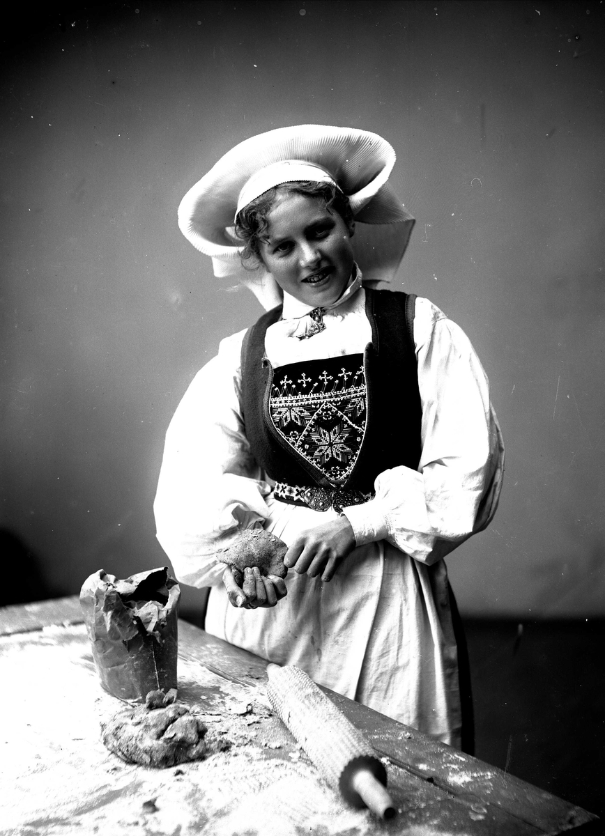 Solveig Lund, 3/4 dels studioportrett av kvinne i drakt og skaut. Hun står bak et bord hvor det ligger kjevle, deig og melpose. I høyre hånd holder hun også deig.