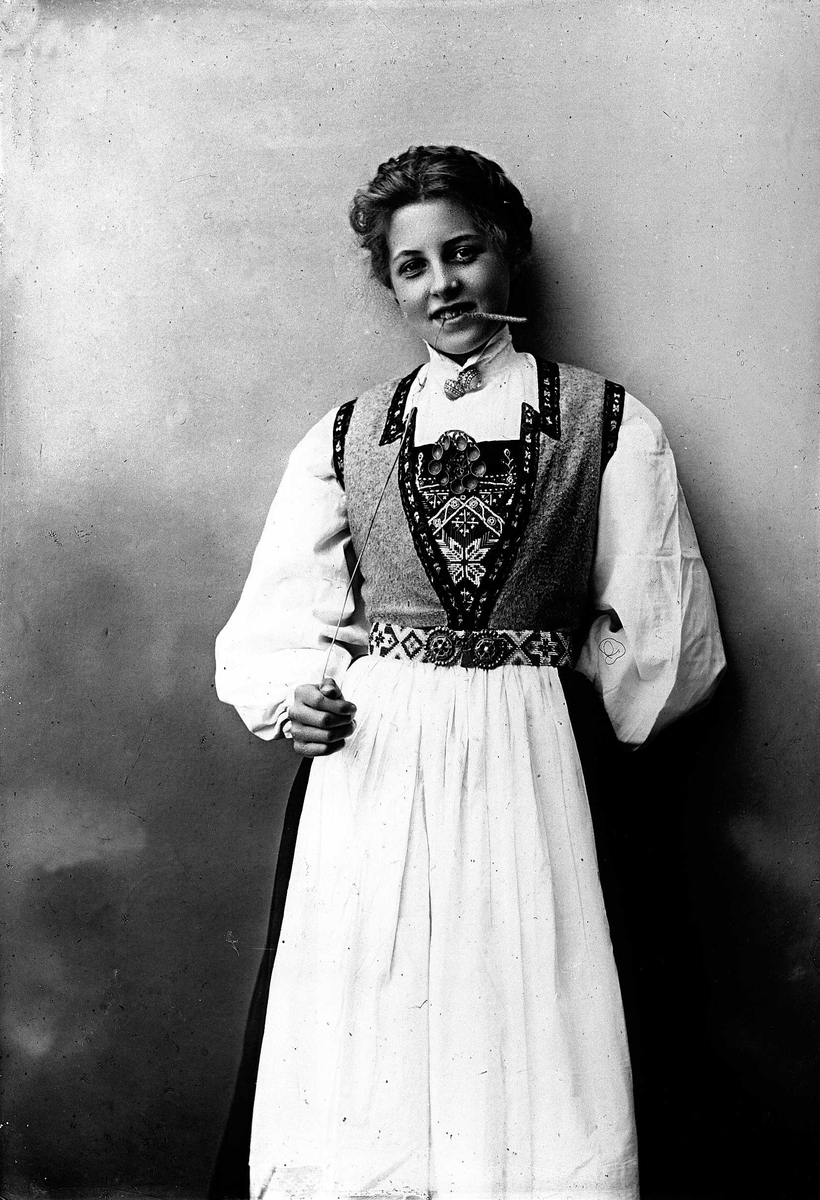 Solveig Lund, 3/4 dels studioportrett av kvinne i drakt, med oppsatt hår. Hun biter på et strå hun holder i høyre hånd. Hun er avfotografert mot en nøytral bakgrunn.