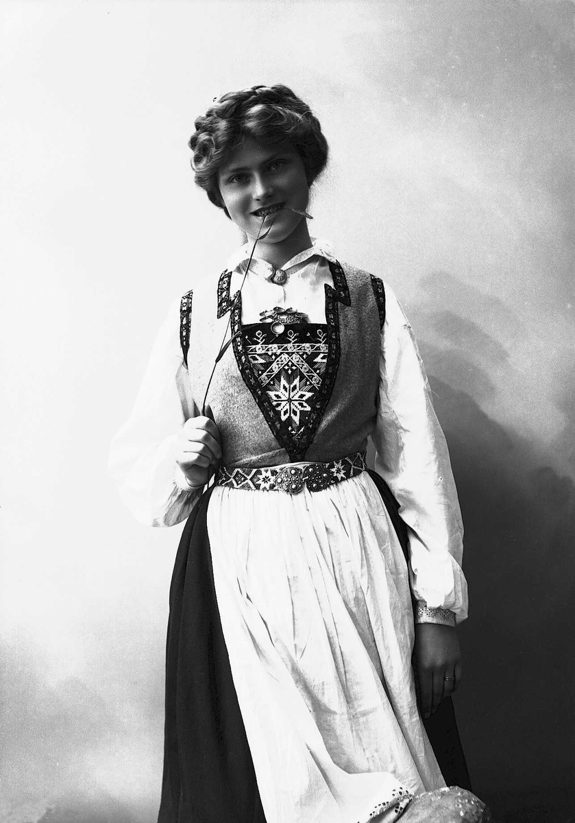 Solveig Lund, 3/4 dels studioportrett av kvinne i drakt, med oppsatt hår. Hun biter på et strå hun holder i høyre hånd. Hun er avfotografert mot en nøytral bakgrunn.