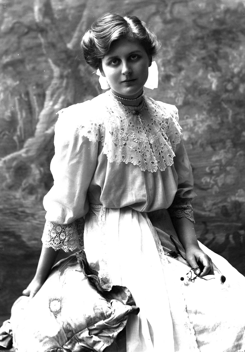 Solveig Lund, 3/4 dels studioportrett av kvinne i drakt, med sløyfe i håret. I venstre hånd holder hun en bukett blomster, mens hun med den høyre støtter seg mot en pute. Bakgrunnen er malt med trær.