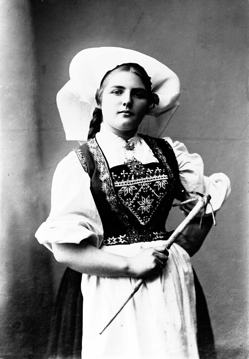 Solveig Lund, 3/4 dels studioportrett av kvinne i drakt, med skaut. Hun holder et redskap i høyre hånd, mens den venstre hviler mot hoften. Bakgrunnen er nøytral.