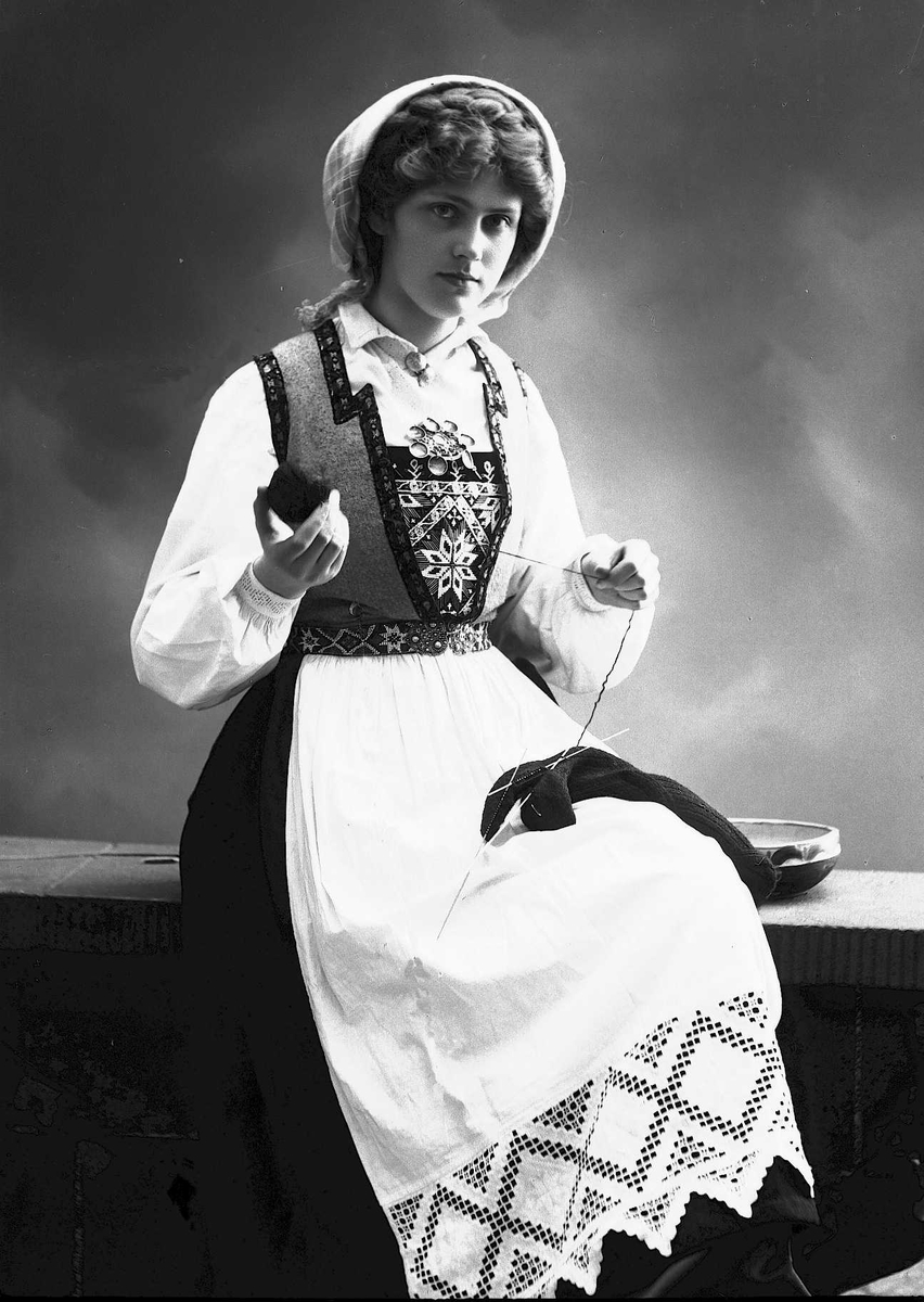 Solveig Lund, 3/4 dels studioportrett av kvinne i drakt og skaut, som sitter på en benk. Hun holder strikketøy i hendene. Bakgrunnen er nøytral.