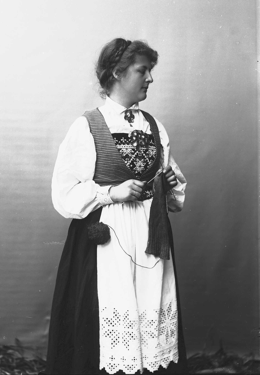 Solveig Lund, 3/4 dels studioportrett av kvinne i drakt, som strikker. Bakgrunnen er nøytral.