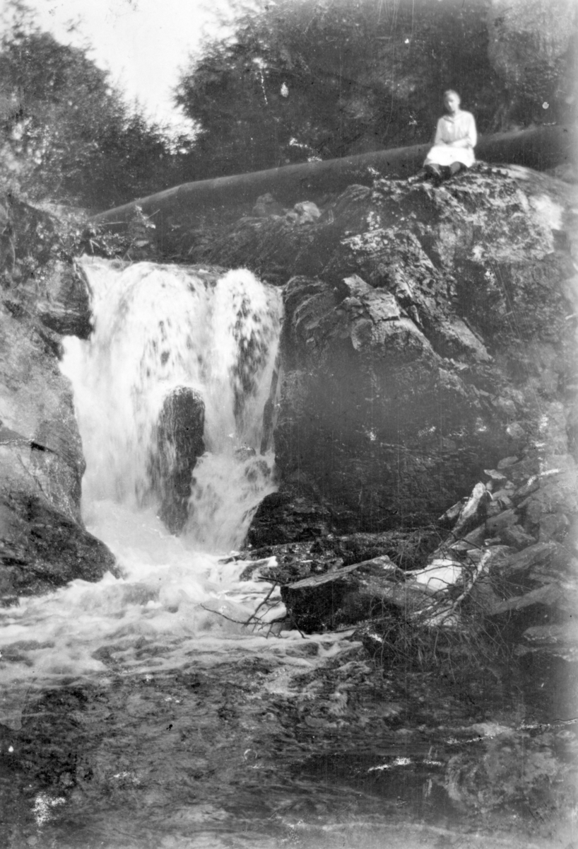 Ukjent, Østre Dystvoll (82/5). Dystvollfossen med stor vannføring. Rørledningen fører vatn til saga. Kvinne sitter ved fossen. Flagstadelva.