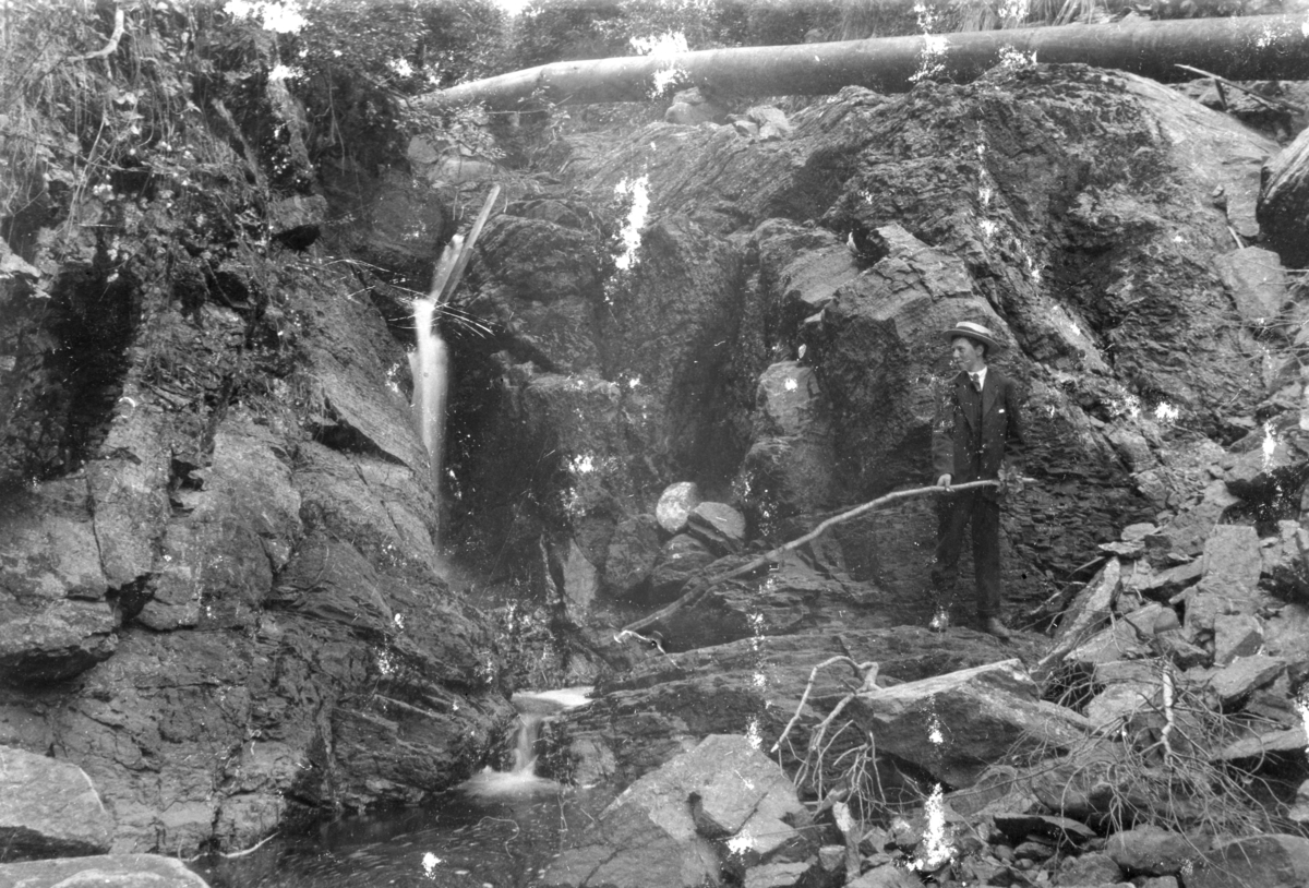 Ukjent, Østre Dystvoll (82/5). Dystvollfossen Vannledningen fører vatn til drift av saga. Harald Olstad fisker i elva under fossen.