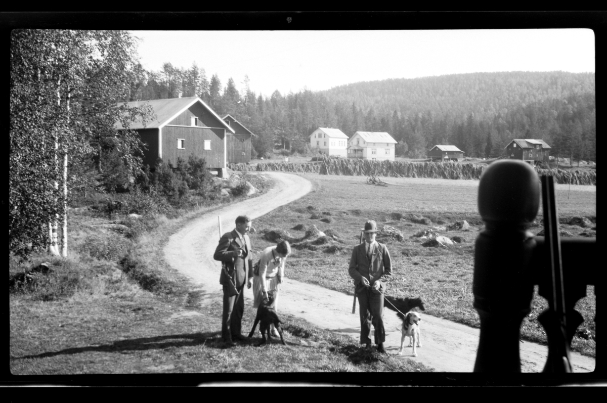 Rolf Sr. Sundt, På vei til eller fra jakt på grusvei mellom gårdsbruk. Fra venstre Johan Buratti med gevær, Hilda Sundt med hund og ukjent mann. Fotografert 1928.