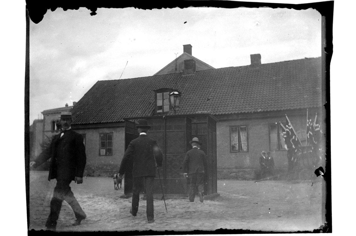 Rolf Sr. Sundt, Menn på vei til og fra urinal på torg i ukjent by. Til høyre flere flagg satt opp i en kjerre. Fotografert 1903-04.