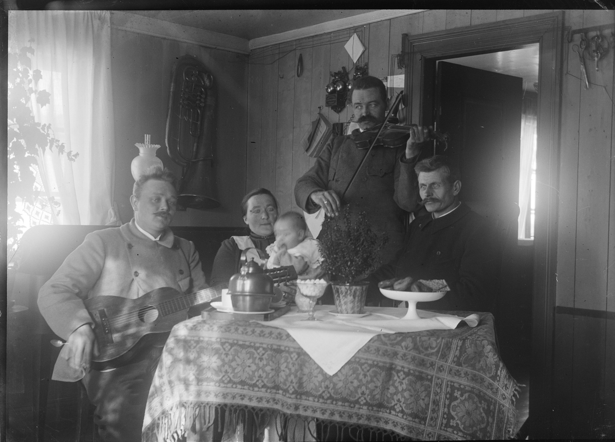 Johannes Larsen Alhaug, Alhaug østre, Furnes. Kaffeslabras, kaffebord. F.v. Johannes Alhaug, Julie Alhaug m/datter Karen Alhaug på fanget, Peder Alhaug (?) m/fele, ukjent. Karen var organist i Furnes kirke i over 50 år. Musikkinstrumenter, gitar, fele. Telefon på veggen i stua.