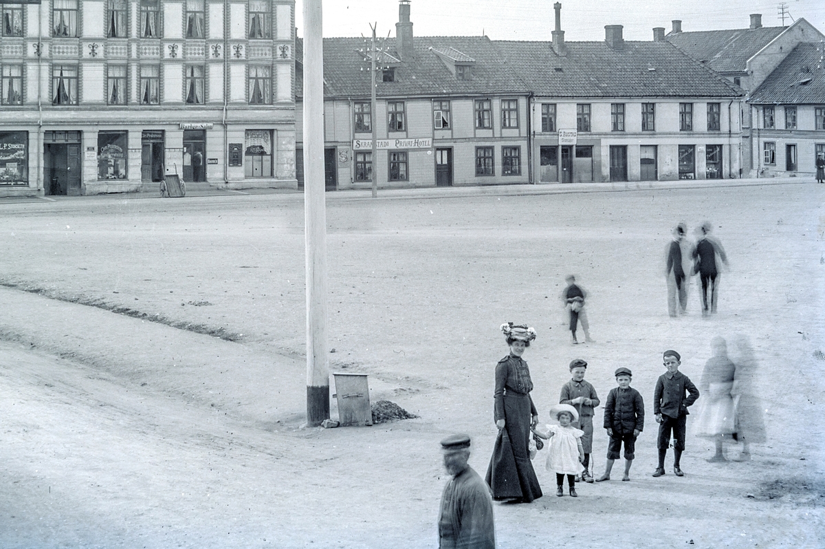 Hans Emil Erichsen, Østre Torg i Hamar. Bebyggelse langs Torggata. Granerudgården med L.P. Storsleth Colonialforretning, Chr. Pedersen Kjødforretning, Skramstad Privathotell, Skomaker S. Rustad. Voksne og barn på torget. Utsnitt.