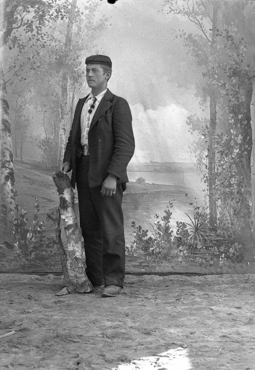 Jakob Jakobsen Engerdal, Ung mann m/cap, dress og lys skjorte (brodert foran). Står ved trestubbe.