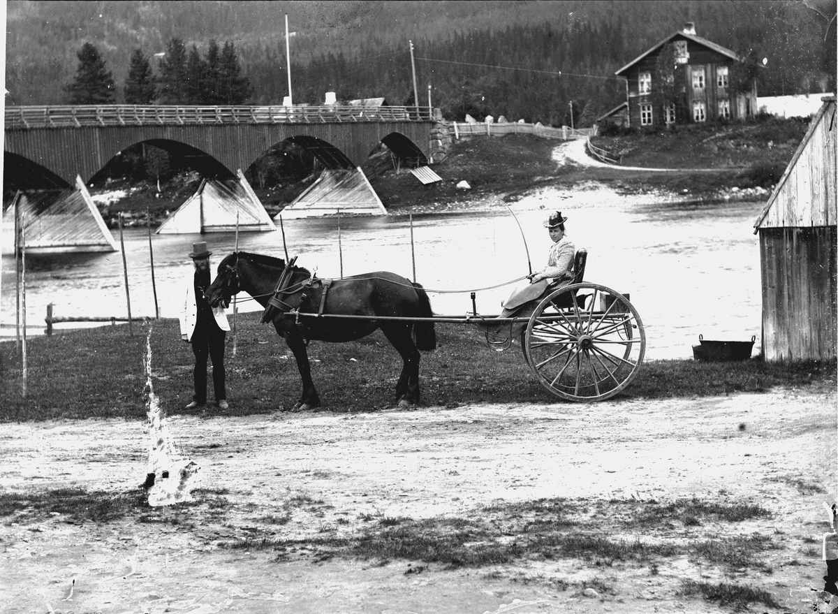 Jakob Jakobsen Engerdal, Nybergsund, bildet er tatt på fotografens egen gårdsplass. Kvinne sitter i en gigg, hest, kjerre, bru, elv.