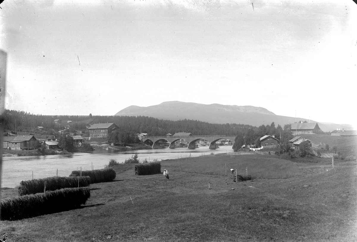 Jakob Jakobsen Engerdal, Nybergsund. Fotografert fra østsida av Trysilelva mot nordvest, Trysilfjellet i bakgrunnen. Brua sto fra 1879-1929. På vestsida av elva ses Nybakk og Nyberg, huset i to etasjer på østsida som ligger delvis skjult bak en høyde er Folkvang.
