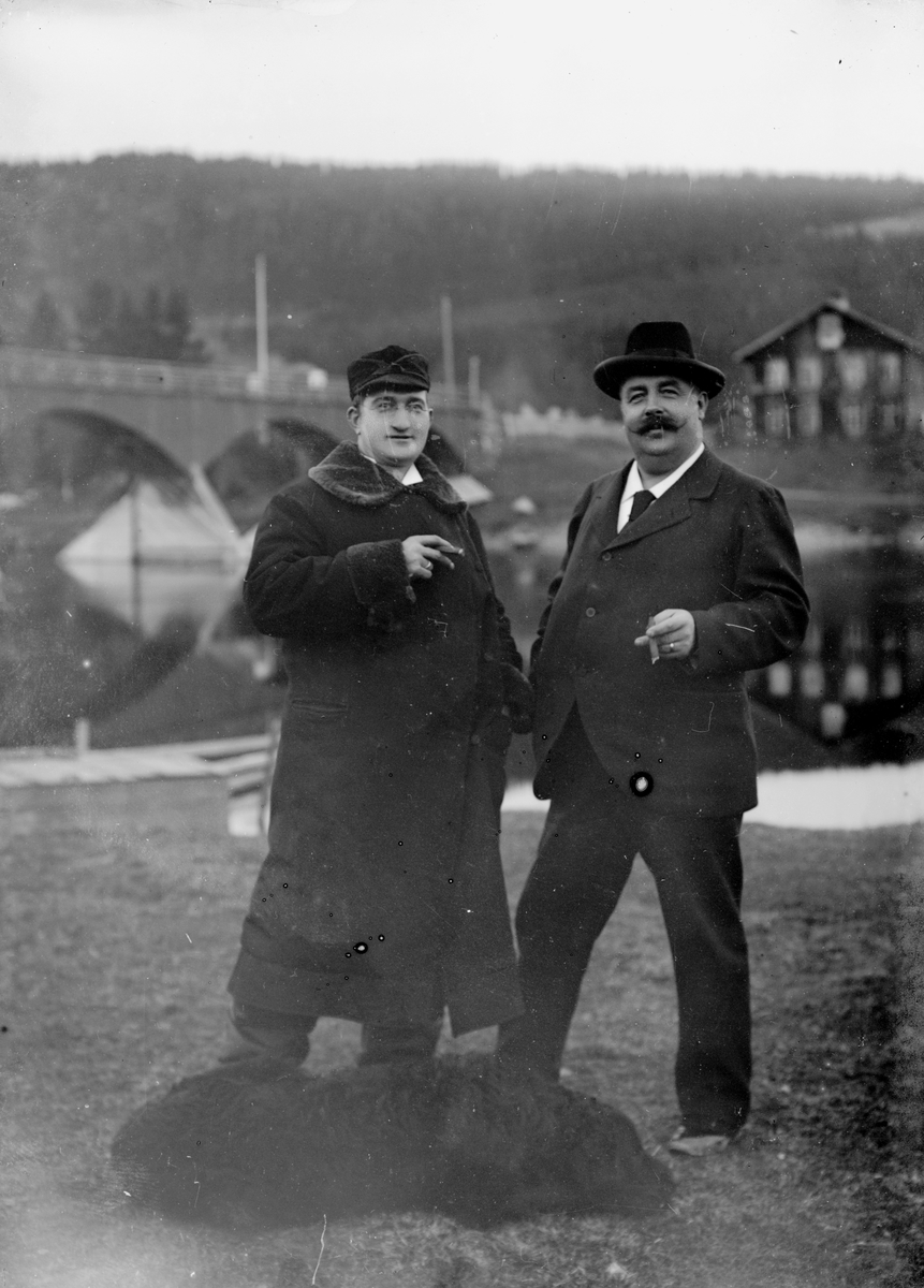 Jakob Jakobsen Engerdal, To menn og en hund foran bru over elva. Til venstre: Doktor (distriktslege i Trysil) Otto Mejlænder (1865-1944). Bildet er tatt på vestsida av Trysilelva, i nærheten av fotograf Engerdals hjem og atelier på Nybakk. Den gamle brua i Nybergsund sto fra 1879-1929. Huset i bakgrunnen er Folkvang, sentr…