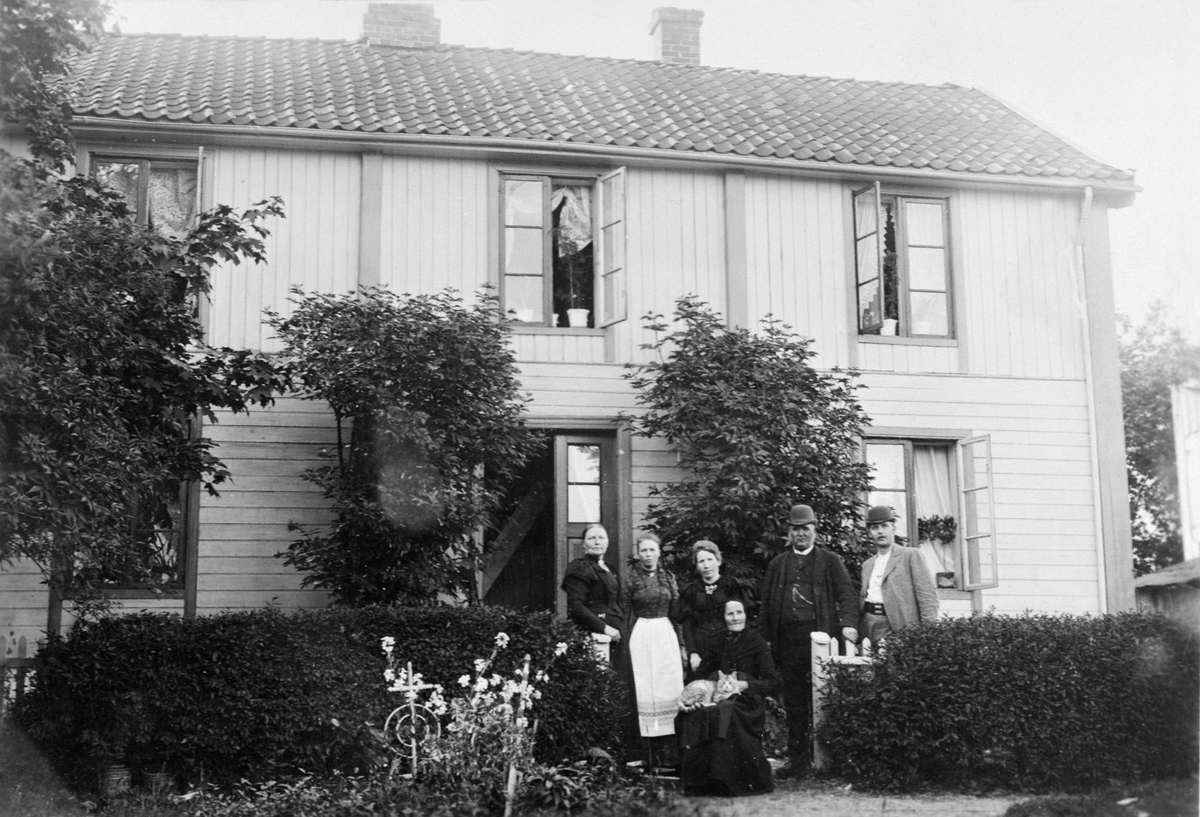 Anno Domkirkeodden, EKSTERIØR, STRANDSTUEN, KIRKEGATA. 11, FAMILIEN FLAGSTAD FORAN HUSET. NR. 2 FRA HØYRE KORPSBØRSEMAKER O. M. FLAGSTAD, MED SØNNEN KAPELLMESTER M. FLAGSTAD OG HUSTRU PIANISTINNEN M. FLAGSTAD NIELSEN PÅ HVER SIDE. YTTERST TIL VENSTRE GAMLE BESTEMOR OLIA FØDT PÅ VALSIG I FURNES SITTER I STOLEN MED KATT…