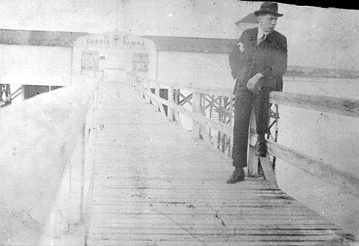 Anno Domkirkeodden, MANN SITTER PÅ REKKVERKET PÅ BRUA TIL HAMAR SJØBAD, BADEHUSET, OVER INNGANGEN STÅR: 1916 HAMAR SJØBAD, MJØSA I BAKGRUNNEN