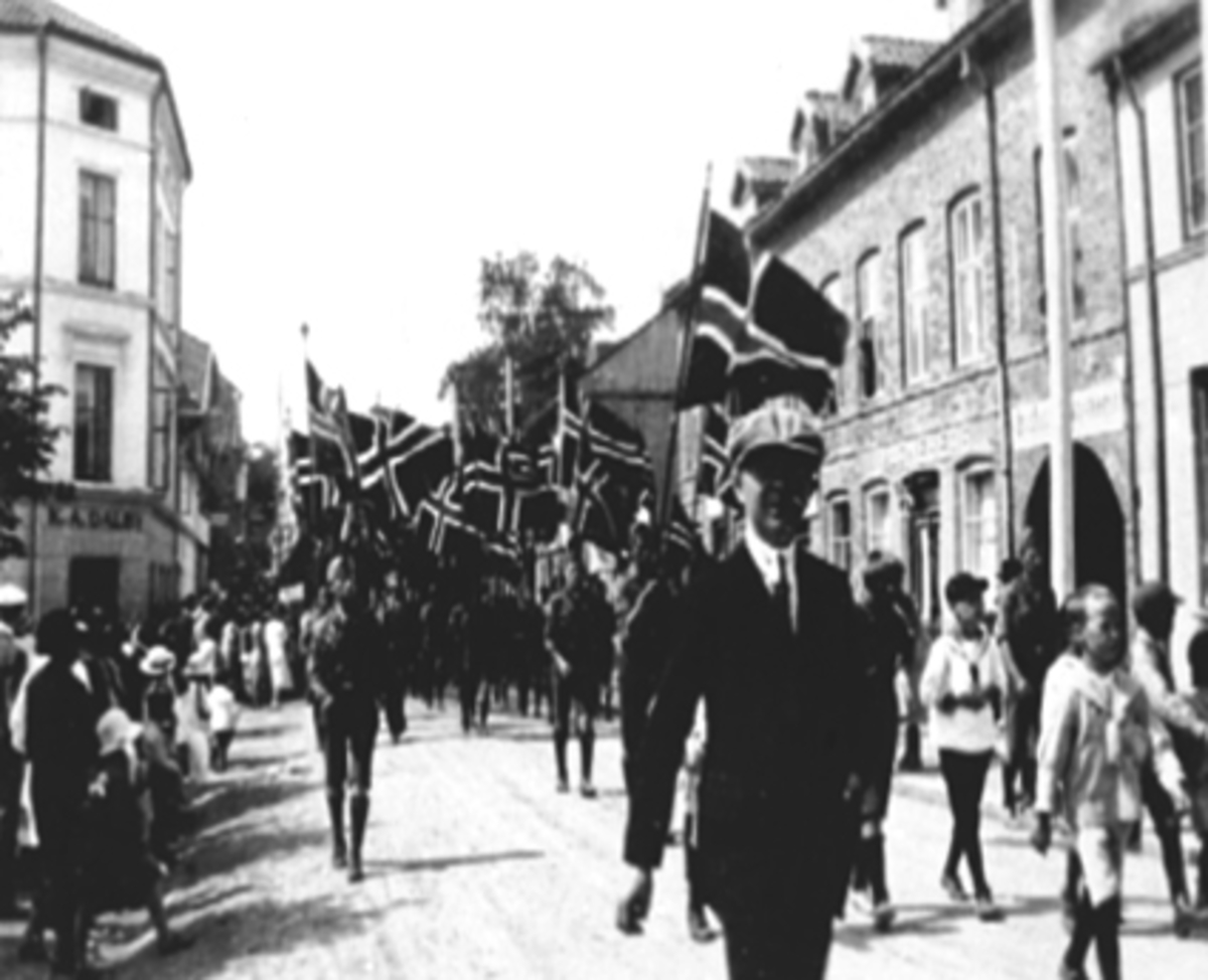 Anno Domkirkeodden, Speidertog marsjerer i Torggata, Stortorget, Hamar. St. Georgsdag 23.04.1925. Gutten t.hh med lyse klær Gustav Norderhaug, gutten forrest Sverre Andersen. Norske flagg.